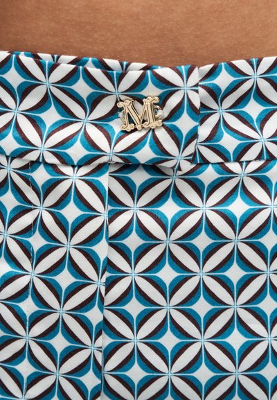Gros plan sur un tissu à motifs géométriques bleu, marron et blanc avec une épingle décorative dorée en forme de lettre « M » attachée près du fond marron.