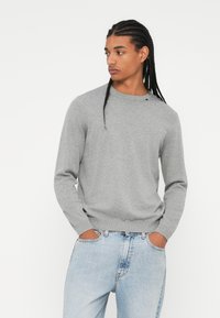 Maglione grigio a maglia con scollatura rotonda e polsini a coste, abbinato a jeans in denim azzurri chiari. Il tessuto appare liscio con dettagli minimi.