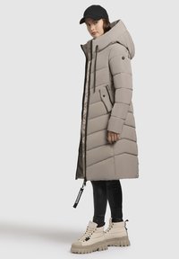 Langer beiger Daunenmantel mit Kapuze, Zickzackmuster und Reißverschluss. Verfügt über eine Reißverschlusstasche und schwarze Akzente. Trägt man mit schwarzen Leggings und klobigen Stiefeln.