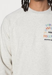 Persoon in een lichtgrijze sweatshirt met meerkleurige geborduurde tekst "Vandaag Morgen Voor Altijd" en het merk "Scotch & Soda."