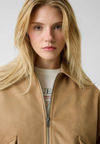 Tan suède jas met ritssluiting en een gladde textuur, voorzien van zijzakken, een hoge kraag en een subtiel geborduurd logo op de borst.