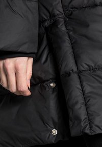Sort pufferjakke med kviltet design, sølvtryklåse og en glat tekstur. Nærbillede af en hånd, der holder stoffet.