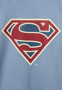 Svetlo modré bavlnené tričko s veľkým, výrazným červeným a žltým logom Supermana. Logo je obkreslené krémovou farbou, čo zdôrazňuje jeho dizajn.