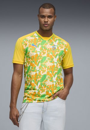 Man draagt een kleurrijk shirt met gele, groene en oranje patronen en korte mouwen, staat met één hand in de zak tegen een grijze achtergrond.