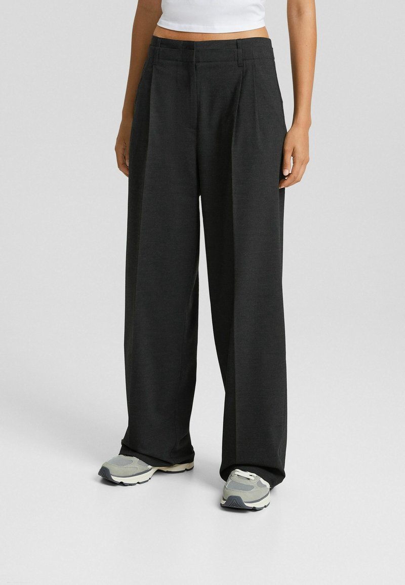 Personne portant un pantalon large noir ample, un top court blanc et des baskets grises, debout sur un fond uni.