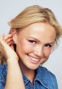 Silver stjärnformade örhängen bärs av en person med ljust blont hår, som ler medan de håller på sitt öra. Denimjackan har synlig sömnad.