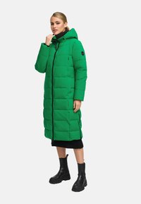 Abrigo acolchado verde con capucha, diseño acolchado, cremallera frontal y bolsillos laterales. Los acentos negros y las botas negras a juego completan el look.