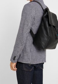 Svart läderryggsäck med en strukturerad yta, med justerbara remmar och ett klaffstängning, bärs över en kavaj med hundtandmönster.