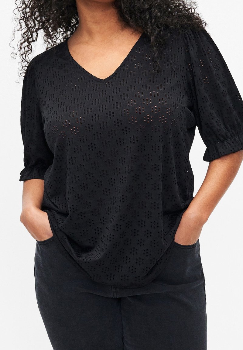 Haut noir tissé avec un col en V, manches courtes bouffantes et motif texturé de petites formes géométriques. Coupe ample avec poches latérales.