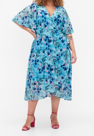 Femme portant une robe portefeuille bleue à motifs floraux avec des manches volantées et des sandales rouges à talons hauts, debout avec une main sur la hanche.