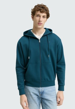 Mėlynas zip-up džemperis su minkšta tekstūra, dviem priekinėm kišenėm, su raišteliu reguliuojama kapučino, ir rūbų apykaklėm, derinamas su šviesiai mėlynomis džinsomis.