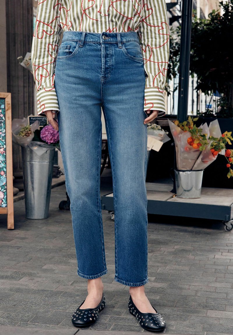 Hoch taillierte blaue Jeans mit geradem Bein, leichter Used-Optik und rohem Saum. Kombiniert mit schwarzen Ballerinas mit silbernen Nieten.