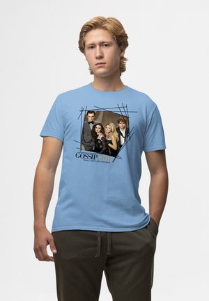 Ung mand iført en lyseblå t-shirt med Gossip Girl-castfoto og teksten "Popularitet har sin pris" foran, stående med den ene hånd i lommen.