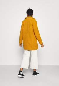 Cappotto giallo textured con un ampio collare, indossato sopra pantaloni bianchi a gamba larga e sneaker alte nere, visto da dietro.
