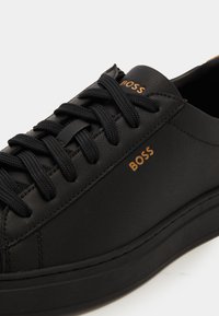 Black low-top sneaker with textured laces and gold "BOSS" logos on tongue and side, detailed stitching on smooth leather upper.