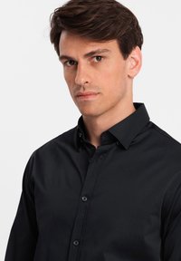 Camicia nera con bottoni, realizzata in tessuto morbido; presenta un colletto, bottoni frontali e maniche lunghe. Design semplice senza motivi visibili.