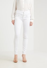 Vita högmidjade jeans med en åtsittande design, som har flera knappar och subtila sömnadsdetaljer, i kombination med en ljus blus.