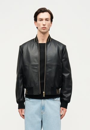 Fiatal férfi fekete bőrbombázó dzsekit visel arany cipzárral és feliratokkal, fekete inget és világoskék farmert, sima háttér előtt áll.