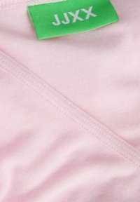 Ružičasti pamučni materijal s glatkom teksturom, s zelenom etiketom označenom "JJXX." Materijal ima čist završetak duž rubova.