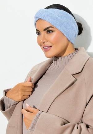 Un serre-tête en laine tricotée bleu clair en matériau moelleux, présentant un design torsadé. Le mannequin porte un manteau beige et un pull côtelé en dessous.