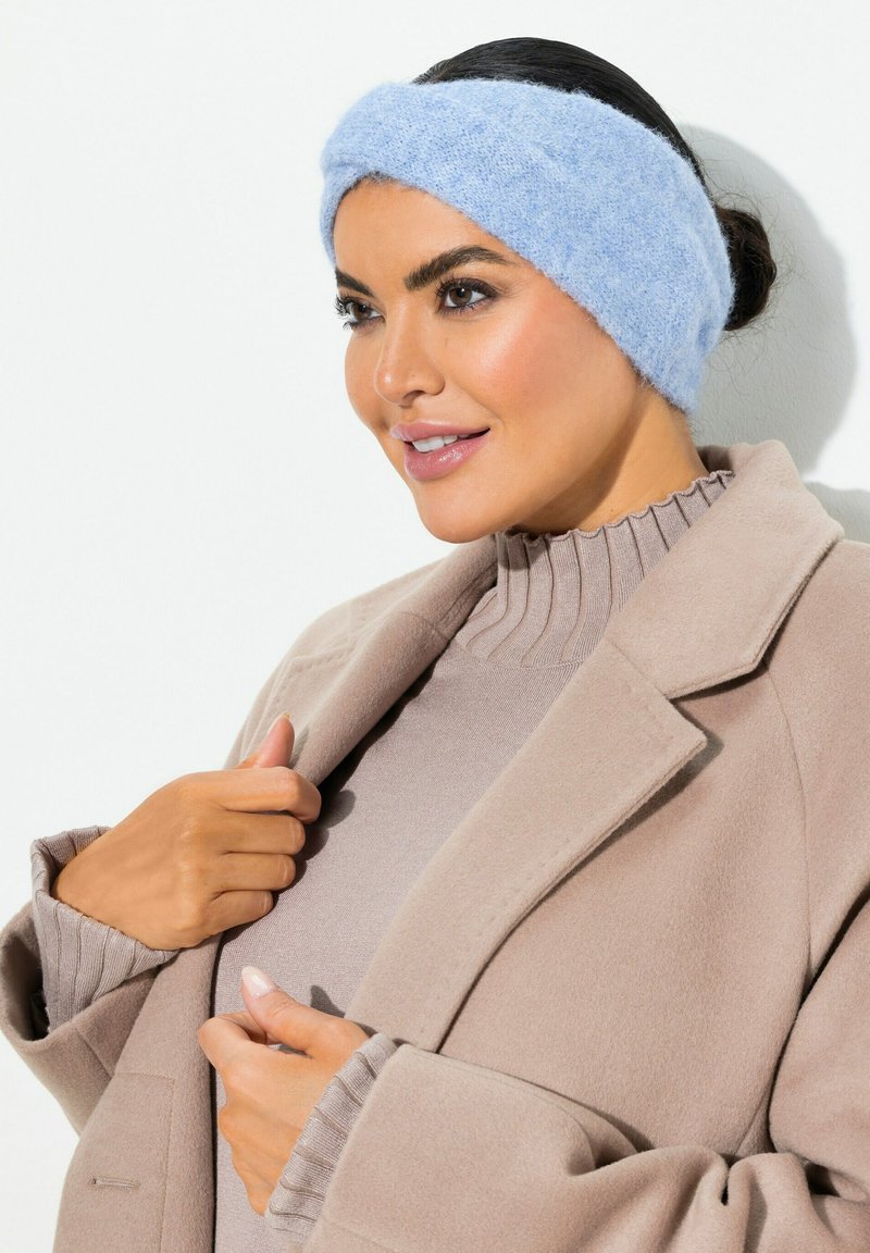 Fascia per capelli a maglia di colore blu chiaro realizzata in materiale soffice, con un design attorcigliato. Il modello indossa un cappotto beige e un maglione a coste sotto.