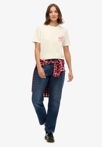 Cremefarvet bomulds t-shirt med et rødt løve grafik på brystet, parret med blå straight-leg jeans og en rød ternet skjorte bundet om taljen. Sorte sko.