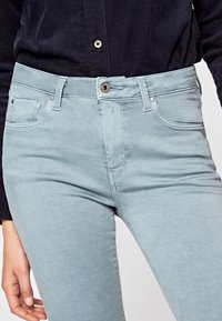 Jeans de color azul claro, hechos de una tela suave, que presentan una cinturilla estándar, bolsillos frontales y detalles en metal plateado. Vista en primer plano de la parte superior.