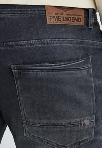 Crni traperice s teksturiranom površinom, s kožnom oznakom na stražnjoj strani označenom "PME LEGEND" i malim logotipom blizu džepa.