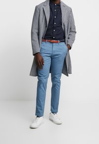 Pánský outfit zahrnující kabát s potiskem pepita, tmavě modrou košili, světle modré kalhoty, hnědý pásek a bílé tenisky.