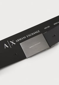 Cinturón de cuero negro con una hebilla de metal con acabado cepillado. La hebilla está grabada con "ARMANI EXCHANGE." Incluye una etiqueta que indica el tamaño y el origen.