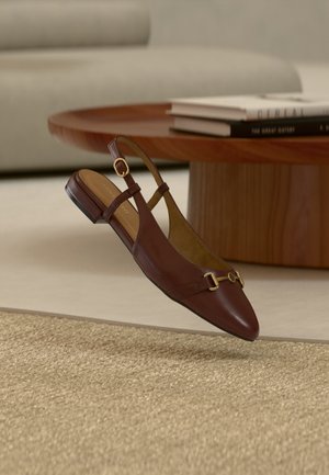 Ruskeat nahkaiset slingback-matalat kengät, joissa kultainen solki, asetettu beigen sävyisen teksturoidun maton päälle lähellä pyöreää puupöytää, jolla on pinottuja kirjoja.