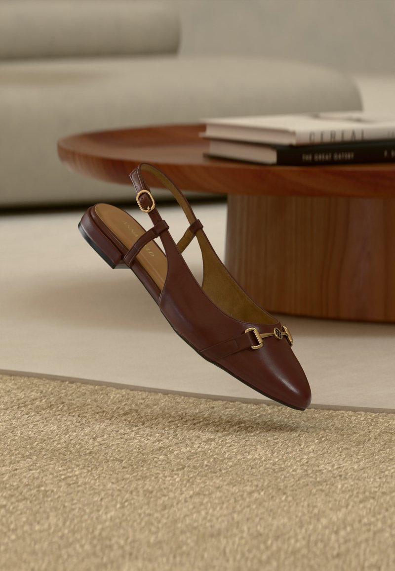 Smeđi kožni slingback ravni cipeli sa zlatnim detaljem kopče, postavljeni na bež teksturirani tepih blizu okruglog drvenog stola sa složenim knjigama.