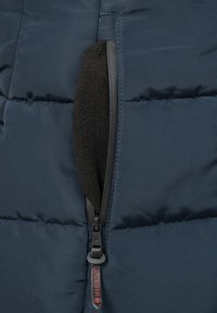 Marinblå pufferjacka med horisontell kviltad design. Har en zip-ficka fodrad med svart fleece. Hållbar dragkedja är synlig.