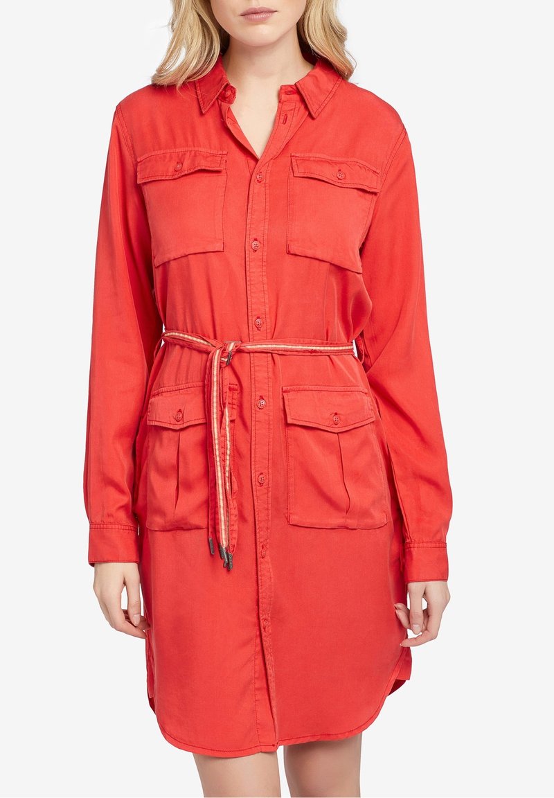 Femme portant une robe chemise rouge vif à manches longues avec des boutons, quatre poches à rabat devant et une fine ceinture nouée à la taille.