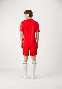 Tricou și pantaloni scurți atletici roșii, din material sintetic, cu un design ajustat. Șosete albe până la genunchi cu dungi negre și ghete albe.