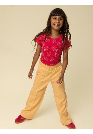 Smiling girl with long dark hair wearing a red strawberry print shirt, beige wide-leg pants, and red sneakers standing on a beige background.