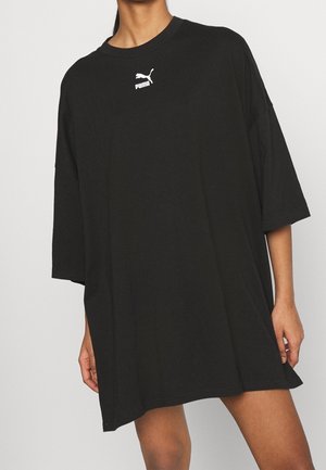 Persoon draagt een oversized zwarte Puma T-shirtjurk met korte mouwen en een logo op de borst, staande tegen een effen achtergrond.