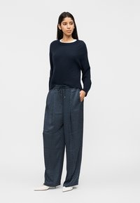 Γυναίκα που στέκεται με τα χέρια στις τσέπες, φοράει μπλε σκούρο πουλόβερ, μπλε σκούρες φαρδιές πουά παντελόνες και λευκά μυτερά παπούτσια.