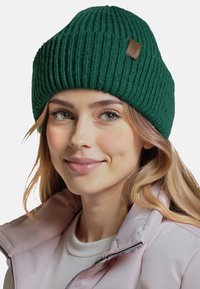 Zielona, żeberkowa czapka beanie z podwiniętym brzegiem i małą skórzaną naszywką, noszona z jasnoróżową kurtką.
