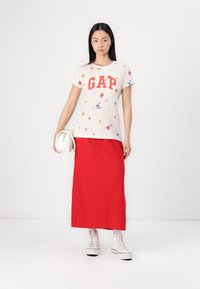 Balta marškinėliai su gėlių raštu ir raudonu "GAP" logotipu, derinami su raudona ilga sijona ir baltomis kanvas bateliais. Modelis laiko baltą rankinę.