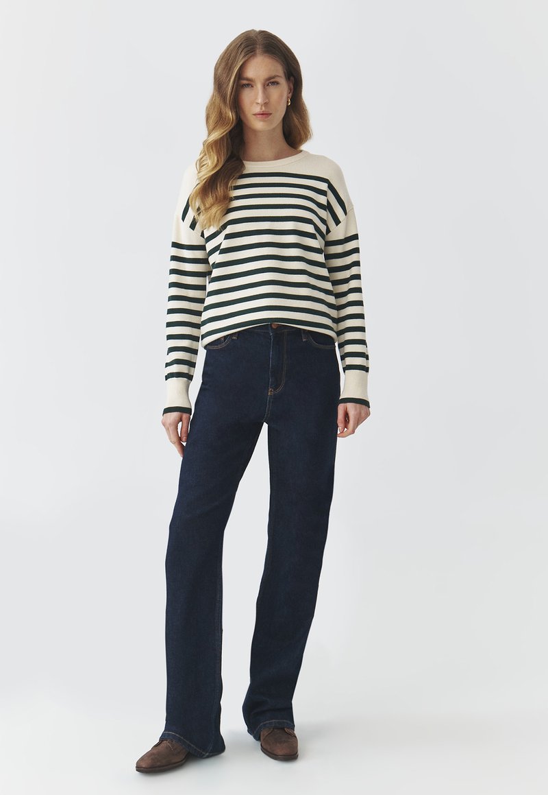 Sweter w paski w kolorze kremowym i ciemnozielonym, z okrągłym dekoltem i obniżonymi ramionami. Połączony z ciemnoniebieskimi dzwonami i brązowymi butami.