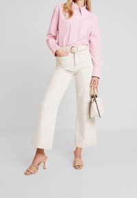 Pruhovaná růžová košile na knoflíky, béžové široké kalhoty, tělové sandály na podpatku a malá bílá taška s dřevěnou rukojetí.