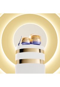 Två Shiseido Vital Perfection krämkrukor, en större och en mindre, på en vit och guld pedestal med en cirkulär guld bakgrund.
