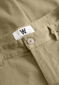 Pantalon en coton beige avec une texture lisse, une fermeture à bouton, et une étiquette blanche avec une marque noire sur la ceinture.