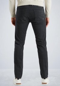 Man som bär slim-fit svarta rutiga byxor och vita sneakers står på ett ljusgrått golv mot en enfärgad bakgrund.