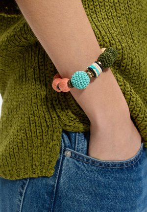 Ręka nosząca masywną, wielokolorową bransoletkę z koralików, odpoczywającą z dłonią w niebieskiej kieszeni dżinsów, widoczny zielony dzianinowy sweter.