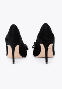 Chaussures à talons hauts en suede noir avec un bout pointu, présentant un talon mince et des accents en métal doré sur la bride de cheville.