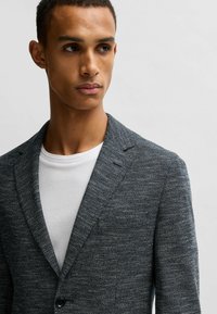 Blazer grigio scuro con texture e un motivo sottile, dotato di rever a punta e chiusura con un solo bottone, indossato sopra una t-shirt bianca con collo tondo.
