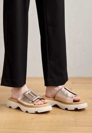 Pieds chaussés de sandales à enfiler métalliques avec de grandes boucles et des semelles blanches épaisses, associés à un pantalon large noir sur un sol en bois.