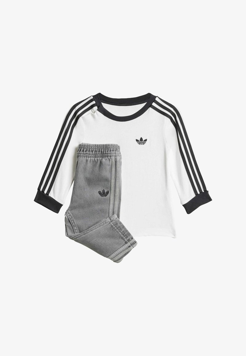 Bílá košile s dlouhým rukávem, na které jsou černé tři pruhy a logo, v kombinaci se šedými tepláky se znakem a odpovídajícími bočními pruhy.